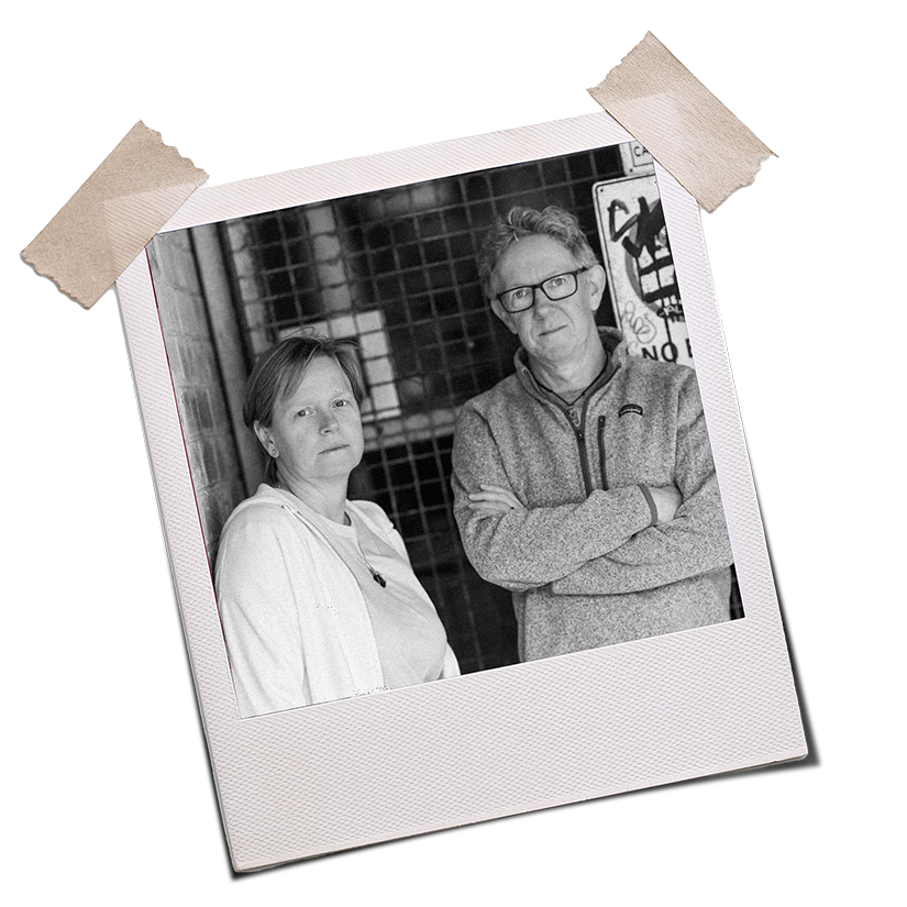 Black and white photograph of authors Bill Duncan and Catherine Lee standing in a dingy alley, in front of a graffitied warning sign. Catherine is leaning against the brick wall and Bill is standing with his arms crossed. Both look very serious.